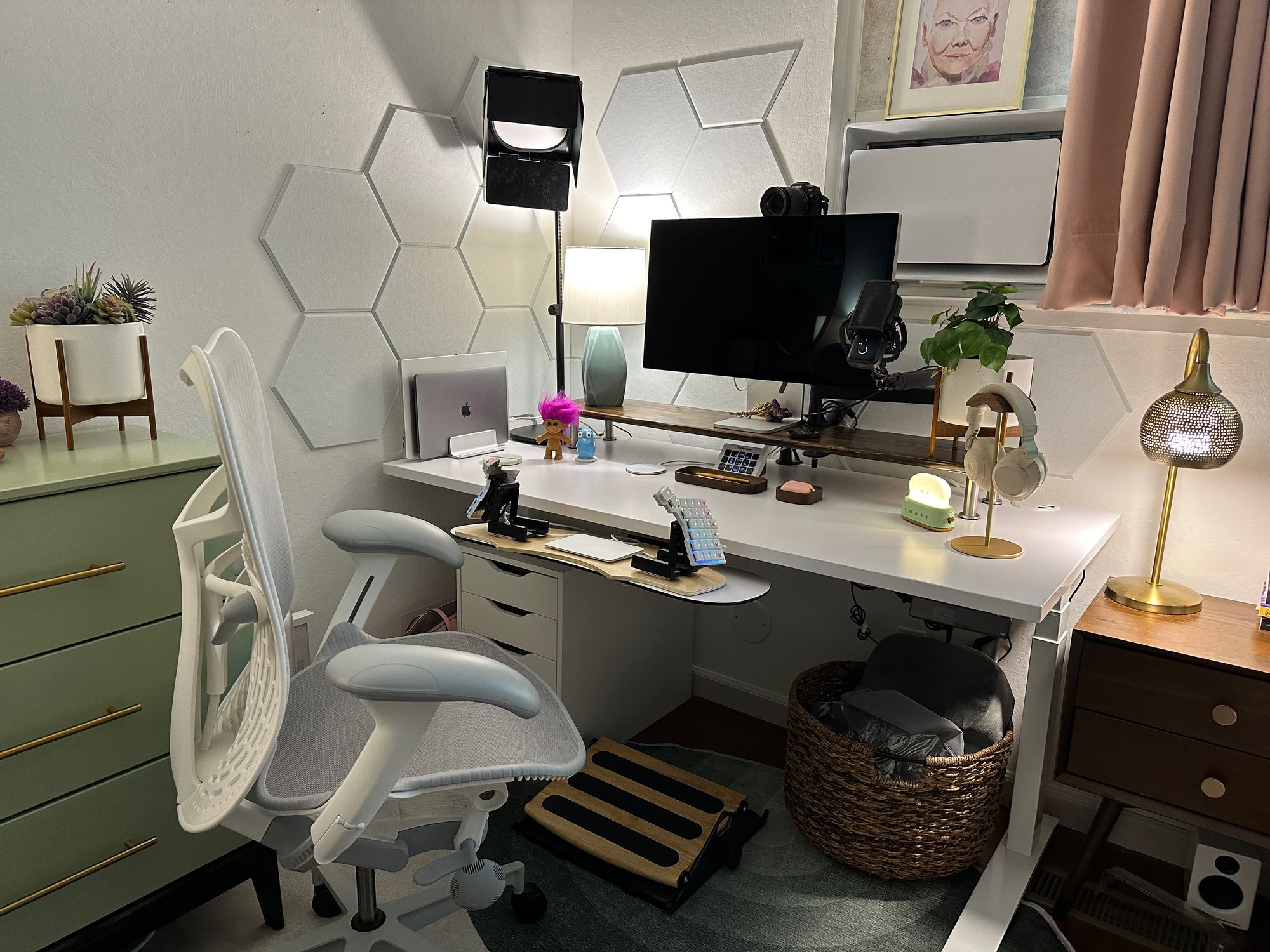 White standing desk with white and grey office chair in front of it, green dresser to the left, and walnut nightstand to the right. On the desk is a monitor on a long dark wood stand along with a turqoise lamp and a leafy plant. Also on the desk are two laptops stood vertically, a few cute figurines, wooden trays for office items, and a gold headphone stand holding white headphones. The desk has a keyboard tray with a split keyboard on it, steeply tented, and a trackpad between the keyboard halves.