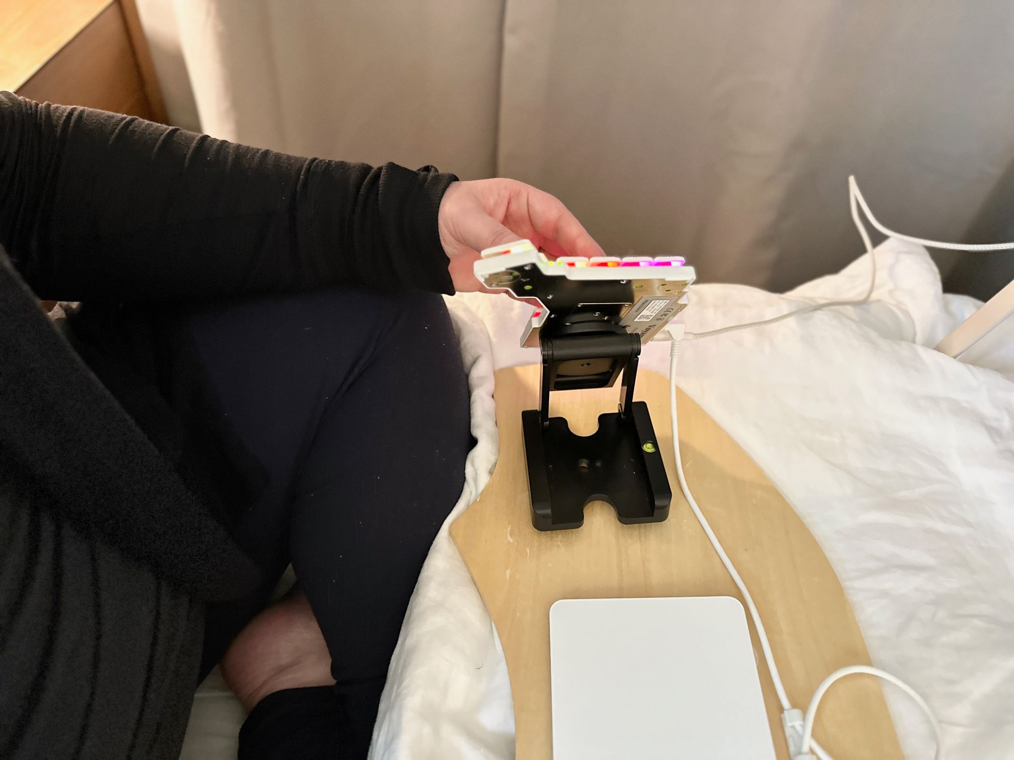 Half of a split keyboard, magnetically attached to a folding stand on a wooden board, which rests on a bed covered with white linen. The stand tents the keyboard very steeply, keeping the wrist of the person using it neutral.